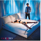 Ad for the Scandal Over the Bed Cross Restraints from CalExotics shows a woman laying on a bed using the product while a man stands next to her.